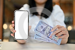 An Asian woman showing a smartphone mockup and carrying a wad of 100 US dollar notes