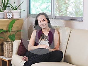 Asian woman sitting on couch in living room with her Chihuahua dog on lap smiling and looking at camera , stay home , social
