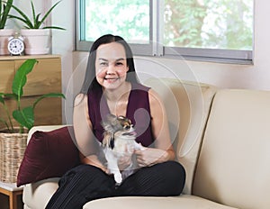 Asian woman sitting on couch in living room with her Chihuahua dog on lap smiling and looking at camera , stay home , social