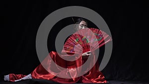 Asian woman sitting on the floor, hides behind a fan and smiles