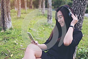 Asian woman sitting and listening to music with headphone and holding smart phone by the tree with green forest background