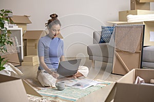 Asian woman sitting in new house using laptop buying decorations online after moving in