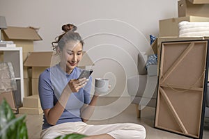 Asian woman sitting in new house using smartphone buying decorations online after moving in