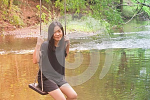 Asian woman sitting relax on wooden swing with water stream and green natural background.