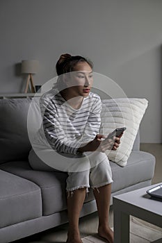 Asian woman sitting on sofa holding phone feeling disappointed, sad, upset