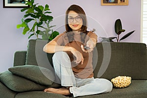 Asian woman sitting on sofa in home living room with bolw of popcorn and using remote control aiming to tv while watching in relax