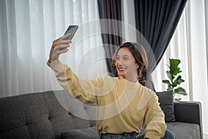 Asian woman sitting on sofa talking with friend via video call and selfie at home