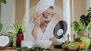 Asian woman sitting at table in white towel and bathrobe, looking at mirror and applying patches