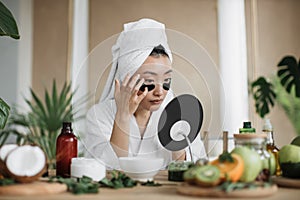 Asian woman sitting at table in white towel and bathrobe, looking at mirror and applying patches