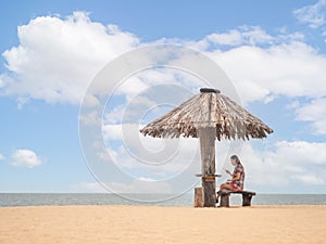 Asian woman sitting under straw umbrella on seashore. female using mobile smart phone on beach. copy space provided.