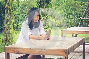 Asian woman sitting on wooden chair at outdoor garden and playing her smartphone.