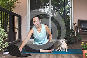 Asian woman  sitting  on yoga mat in balcony  with Chihuahua dog, using  computer laptop. Yoga exercise training online