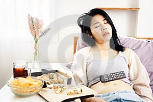 Asian woman sleeping in bed after eating pizza, potato chips and soda with Tv remote on her fat belly  overeating junk food