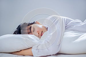 Asian woman sleeping well in bed hugging soft white pillow with blanket on white sheets in bedroom