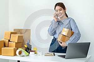 Asian woman small business owner holding parcel of box and talking with customer on smartphone to receive order