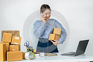 Asian woman small business owner holding parcel of box and talking with customer on smartphone to receive order