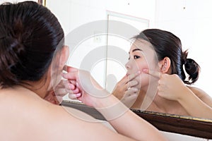 Asian woman squeezing pimples on her face while looking at reflection in mirror.