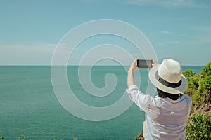 Asian woman standing relax on cliff nearly seashore, She taking photograph seascape view with smartphone.