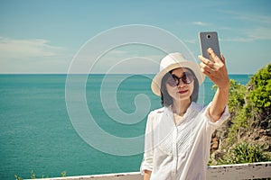 Asian woman standing relax on cliff nearly seashore, She taking selfie with smartphone.