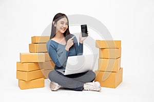 Asian woman startup small business freelance holding parcel box, mobile phone and computer laptop and sitting on floor