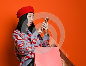 Asian woman in a stylish beret and sweater, with a colorful shopping bag looks into her mobile phone and something is surprised