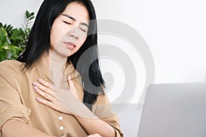 Asian woman suffering from heartache hand holding her chest pain while working at work sitting at desk try to breath
