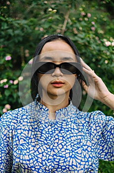 an Asian woman with sunglasses and a bandanna posing elegantly in a blue patterned shirt in front of flowers