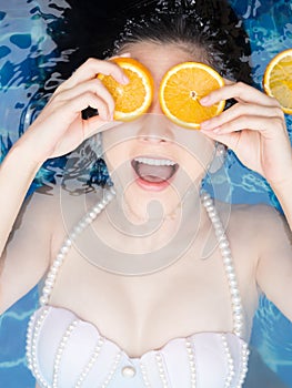 Asian woman is swimming with a slice of orange floating above the water in a blue pool and enjoying a hot summer day.