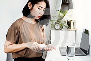 Asian woman taking some painkiller pills from a bottle while working on computer at desk