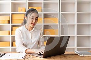 Asian woman talking on smartphone with customer, keying commands with laptop, small business, e-commerce