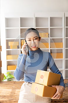 Asian woman talking on smartphone with customer, keying commands with laptop, small business, e-commerce