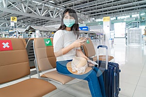 Asian woman tourist wearing mask using mobile phone searching airline flight status and sitting social distancing chair in airport