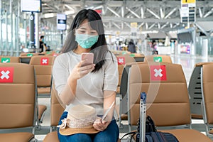 Asian woman tourist wearing mask using mobile phone searching airline flight status and sitting social distancing chair in airport