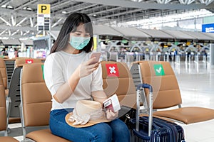 Asian woman tourist wearing mask using mobile phone searching airline flight status and sitting social distancing chair in airport