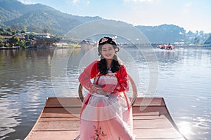 Asian woman tourist wearing red Hanfu ancient Chinese traditional ethnic dress sitting on wooden boat at reservoir of Ban Rak Thai