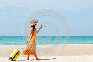Asian women traveler holding yellow luggage walking and check in smart phone on the beach photo
