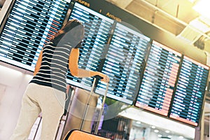 Asian woman traveler looking at flight information screen in an airport, holding suitcase, travel or time concept