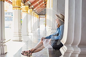 An Asian woman traveler in white dress and jean jacket and backpack, sitting on temple pavilion floor at Wat Phra Kaew Emeral