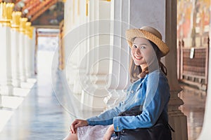 An Asian woman traveler in white dress and jean jacket under straw hat sitting on floor, smile on face and backpack