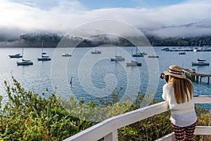 Asian woman traveller taking picture of Blue sky, white clouds, mist & aquamarine seas at Akaroa Harbor. Anchored are fishing