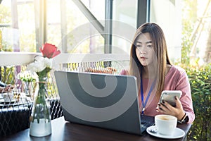 Asian woman using laptop computer and smart phone with white coffee cup on table in cafe