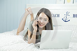 Asian woman using laptop and speak on phone while lying on her bed.
