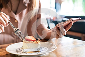 Asian woman using mobile phone while eating cake on wooden table in cafe. Relaxation, technology and lifestyle concept. Close up