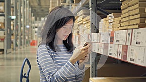 Asian woman using a smart phone or shopping on shelves at large warehouse retail store industry. Rack of furniture and home
