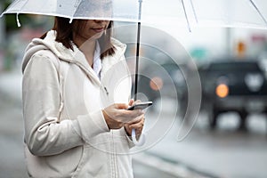 Asian woman is using on smartphone, checking social media network and holding umbrella while waiting taxi on the city street