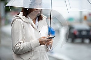 Asian woman is using on smartphone, checking social media network and holding umbrella while waiting taxi on the city street