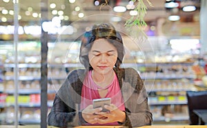 Asian woman using smartphone on counter at supermarket with glass window reflection