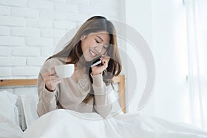 Asian woman using the smartphone on her bed while holding cup of coffee in the morning.