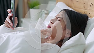 Asian woman waking up in the morning and using smartphone in bed feeling happy.