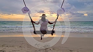 Asian woman watching sunset on the beach at a swing under a palm tree